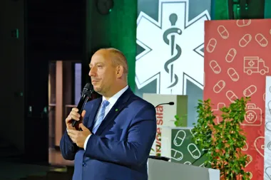 Mężczyzna w granatowym garniturze przemawia do mikrofonu podczas wydarzenia medycznego, w tle symbol ratownictwa medycznego i grafiki z pigułkami.