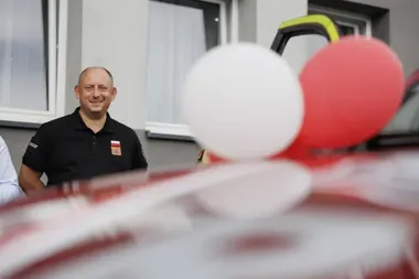 Mężczyzna w czarnej koszulce z flagą Polski i emblematem stoi przed budynkiem, na pierwszym planie balony i wstęgi.