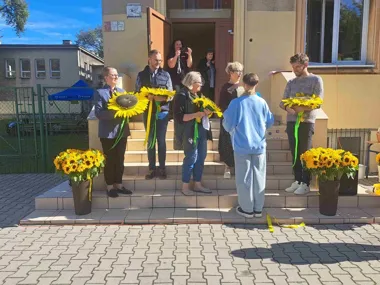 Chłopiec wręcza duże słoneczniki grupie dorosłych stojących na schodach przed budynkiem w słoneczny dzień.