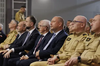 Mężczyźni w mundurach służb ratowniczych i w garniturach siedzą w rzędach podczas oficjalnego spotkania lub uroczystości w pomieszczeniu.