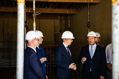 Mężczyźni w garniturach i białych kaskach ochronnych rozmawiają na budowie wewnątrz nieukończonego budynku.