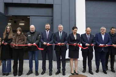 Grupa osób oficjalnie przecina czerwoną wstęgę podczas uroczystego otwarcia nowego obiektu przed budynkiem przemysłowym.