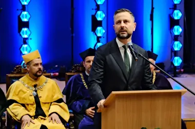 Mężczyzna w garniturze przemawia z drewnianego pulpitu podczas uroczystej akademickiej ceremonii, w tle siedzą osoby w togach i czapkach.