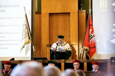 Uroczystość w auli AGH w Krakowie: mężczyzna w togi rektorskiej przemawia z pulpitu, obok sztandary uczelni, na pierwszym planie osoby w biretach.