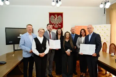 Grupa osób w sali urzędowej z polskim godłem na ścianie pozuje po podpisaniu dokumentów, które trzymają dwaj mężczyźni.