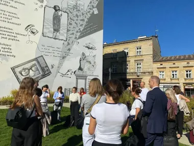 Grupa osób stoi na trawniku przed wielkim czarno-białym muralem z ilustracjami i tekstem, w tle kamienica.