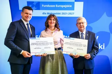 Trzy osoby stoją przed niebieskim tłem z napisem „Fundusze Europejskie dla Małopolski 2021-2027” i trzymają duże czeki symbolizujące dofinansowanie projektów re