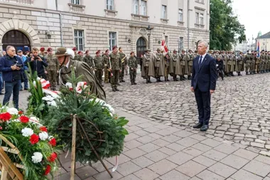 Mężczyzna w garniturze stoi przed wieńcem złożonym na brukowanej płycie podczas uroczystości z udziałem żołnierzy i orkiestry wojskowej przed historycznym budyn