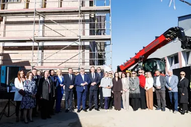 Grupa osób pozuje do zdjęcia na placu budowy przed budynkiem otoczonym rusztowaniem, obok stoi czerwona ładowarka teleskopowa Manitou.