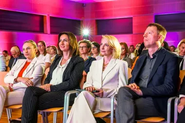 Publiczność siedzi na krzesłach w sali oświetlonej fioletowym i różowym światłem, słuchając wydarzenia konferencyjnego.