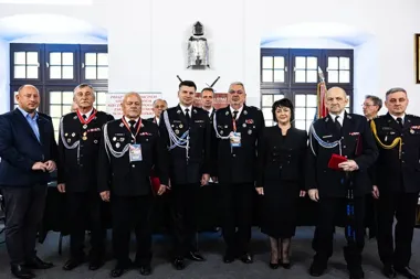 Grupa mężczyzn w mundurach strażackich, kobieta w czerni i mężczyzna w garniturze pozują do zdjęcia grupowego w pomieszczeniu z oknami i sztandarem.