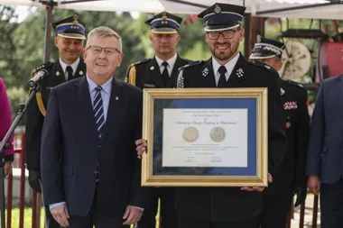 Mężczyzna w garniturze i funkcjonariusz straży pożarnej w mundurze trzymają wspólnie oprawiony dyplom podczas uroczystości na wolnym powietrzu.