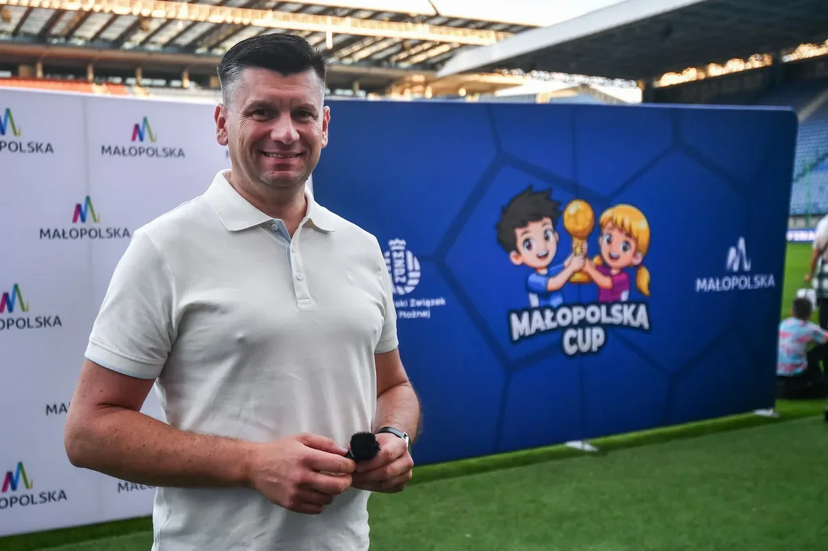 Mężczyzna w białej koszulce polo stoi na murawie stadionu przed ścianką reklamową Małopolska Cup.